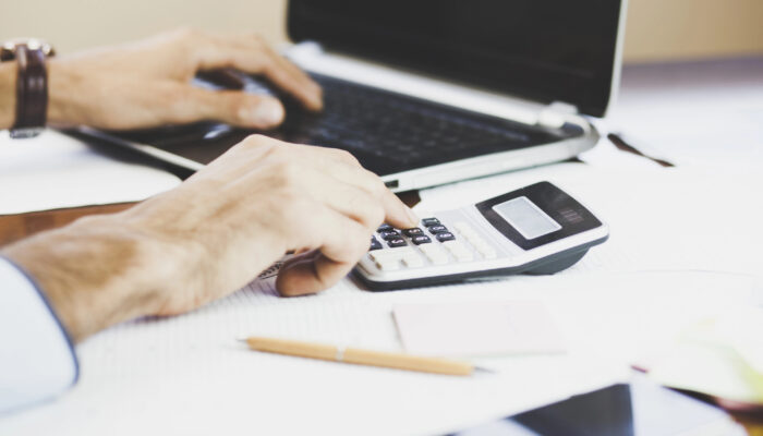 Close up of a businessman typing on a calculator, with a laptop open next to it.